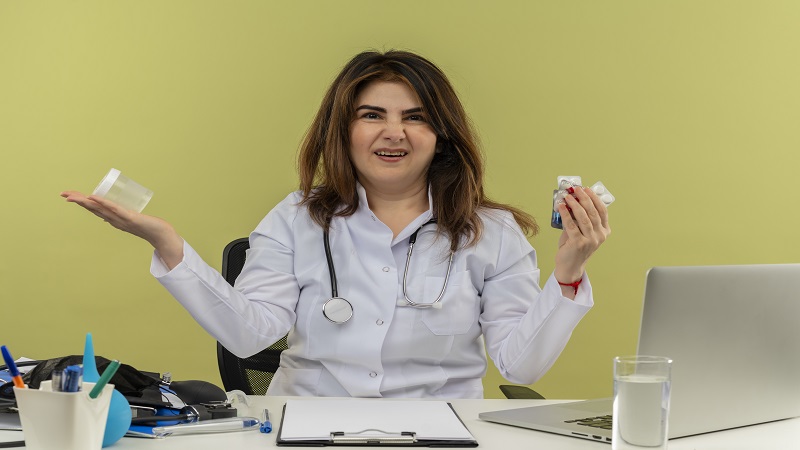 annoyed middle-aged female doctor wearing medical robe and stethoscope sitting at desk with medical tools and laptop holding medical drugs and beaker isolated on olive green background